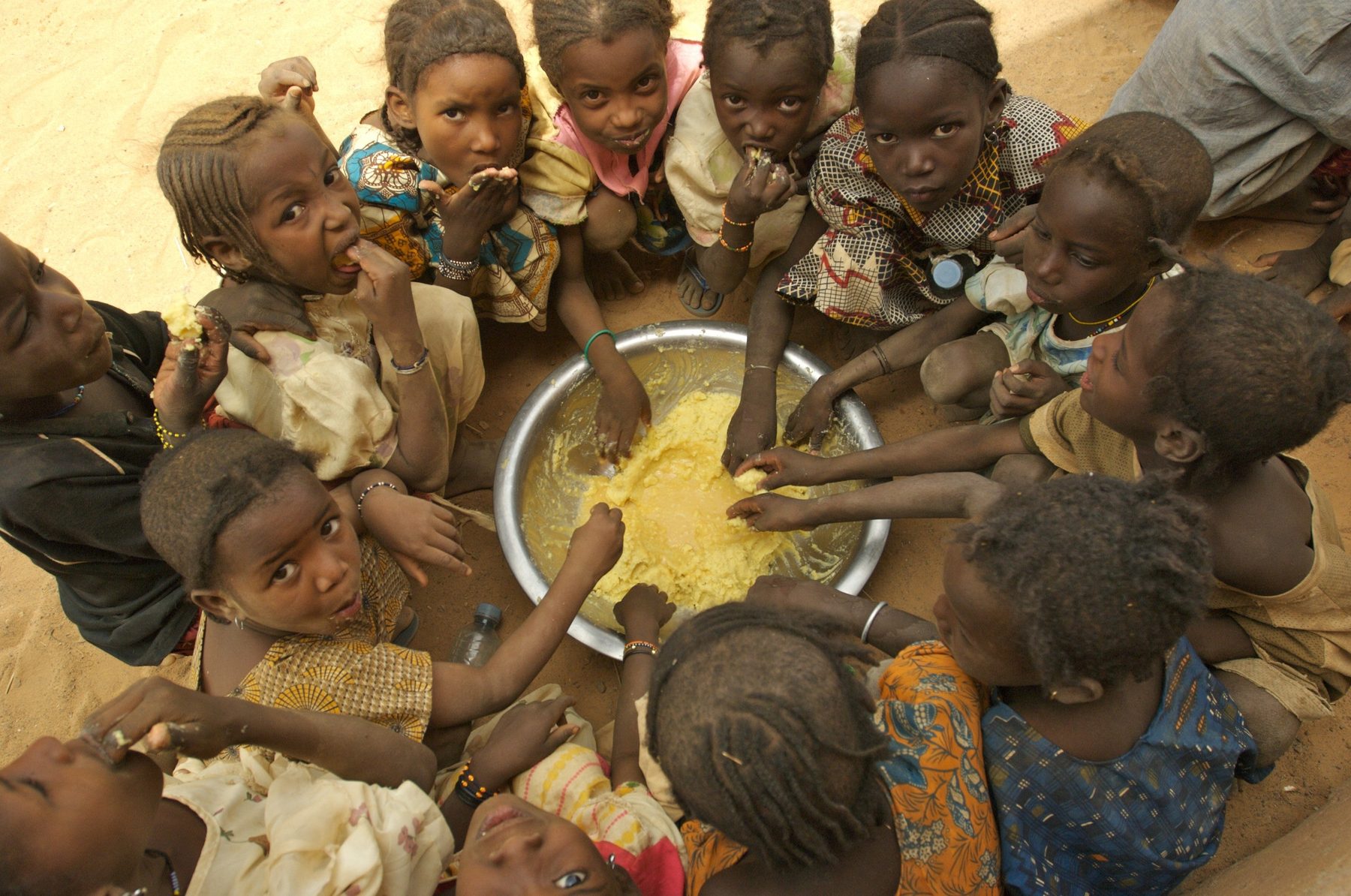 A group of children sitting on the sand, sharing a meal from one large bowl